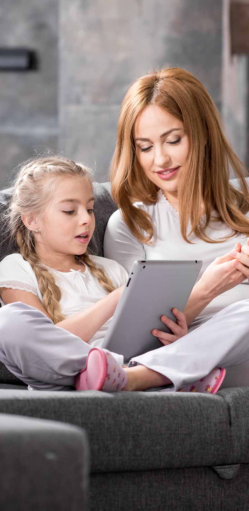 Mother and daughter with red hair sitting on a grey couch looking at a tablet
